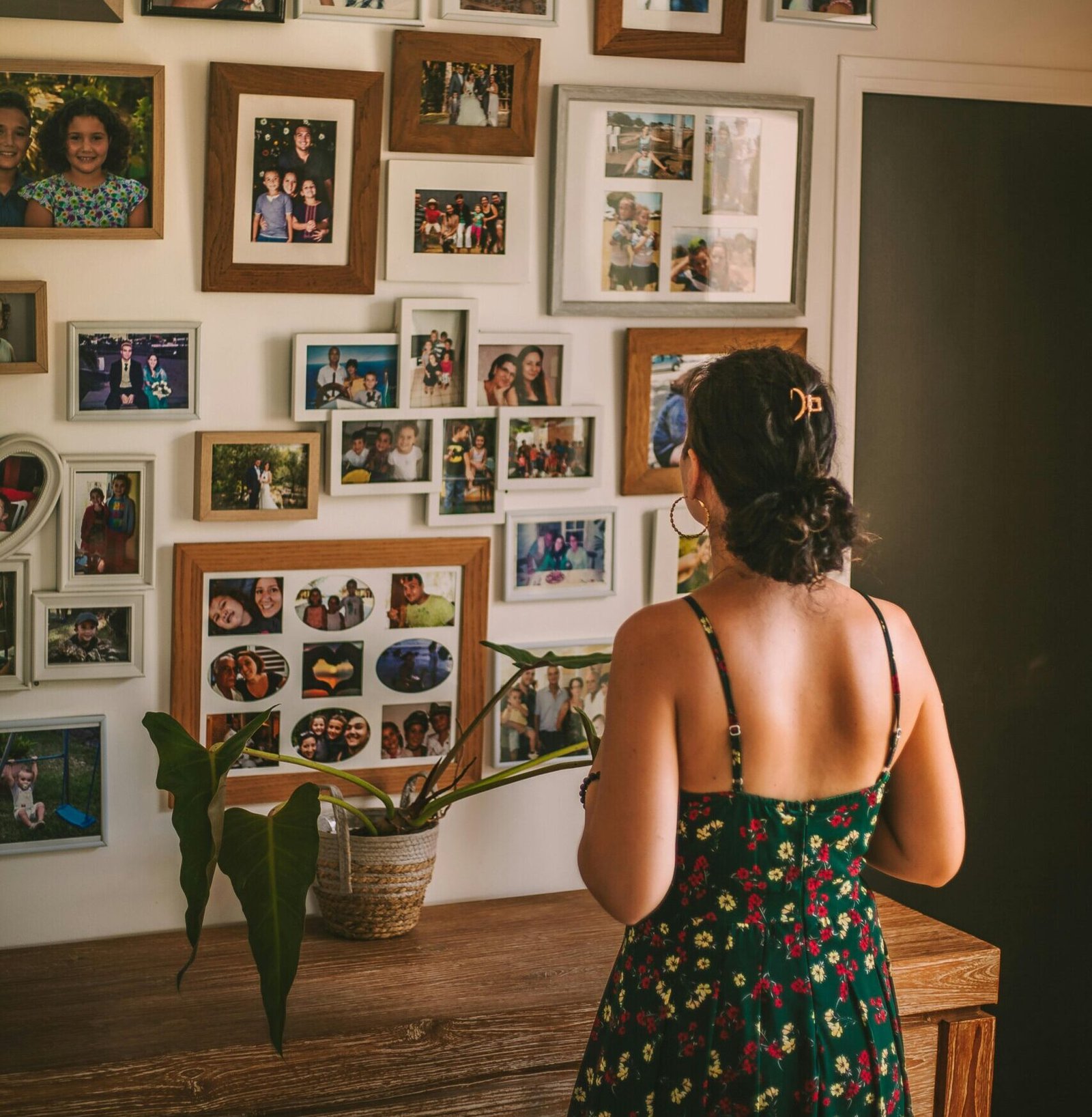 A woman in a floral dress admires family photo frames on a living room wall.